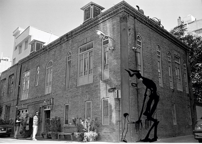 A black and white street mural depicting a tall, spindly, shadowy figure interacting with a smaller human character on a brick building corner.