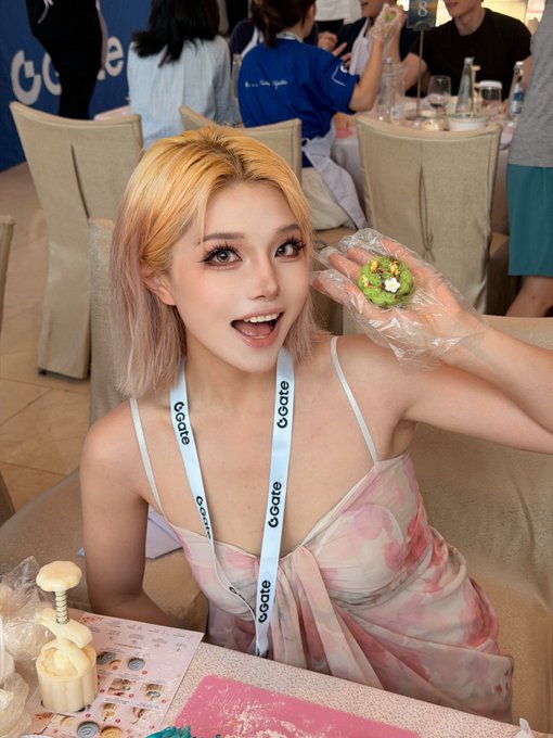 A young woman with blonde hair smiling and holding up a small green treat in a plastic-gloved hand, wearing a lanyard with 'GOGATE' written on it.