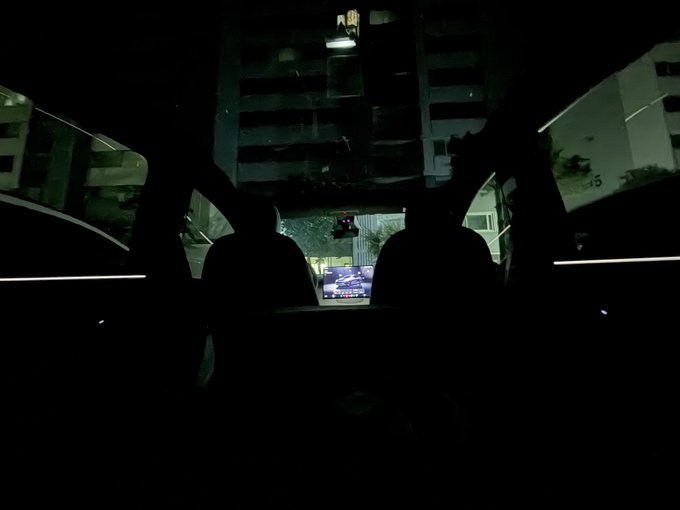 The image shows the dark interior of a Tesla Model Y at night, looking forward through the windshield at an illuminated building and a red traffic light visible on the dashboard display. This scene visually represents the owner's humorous decision to "camp out" in their financed Model Y following the Dogecoin crash, capturing the bleak mood of someone seeking refuge in their car.