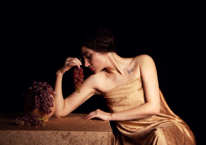 A dramatic, low-key lit portrait of a woman in a gold dress leaning on a stone table, holding a cluster of red grapes near her face with a clay pot of grapes beside her against a black background.