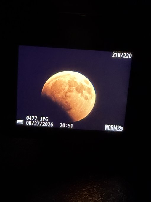 A photograph of a camera screen displaying an image of a partially shadowed moon, with the text '0477.JPG', '08/27/2026 20:51', '218/220', and 'NORM'.