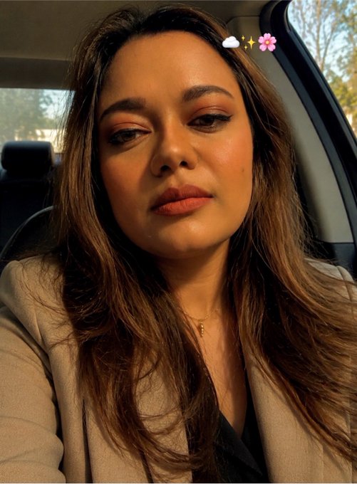 A selfie-style photo of a young woman with long brown hair and makeup, sitting in a car, with a small flower and sparkle emoji filter visible near her hair.