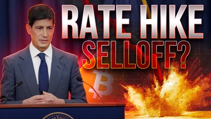 A news anchor at a podium is featured alongside bold text reading "RATE HIKE SELL OFF?" set against a fiery explosion backdrop with a faint Bitcoin logo, visually reinforcing the post's warning about rate hike fears causing a crypto sell-off. The dramatic imagery aligns with the video's alarmist headline about dumping cryptocurrencies like $BTC, $ETH, $SOL, $XRP, and $VIRTUAL.