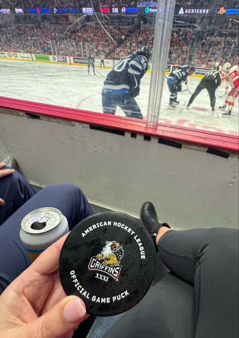 A first-person photo at a hockey game showing a hand holding a black American Hockey League official game puck for the Griffins (game XXXI), with players on the ice visible through the glass in the background.