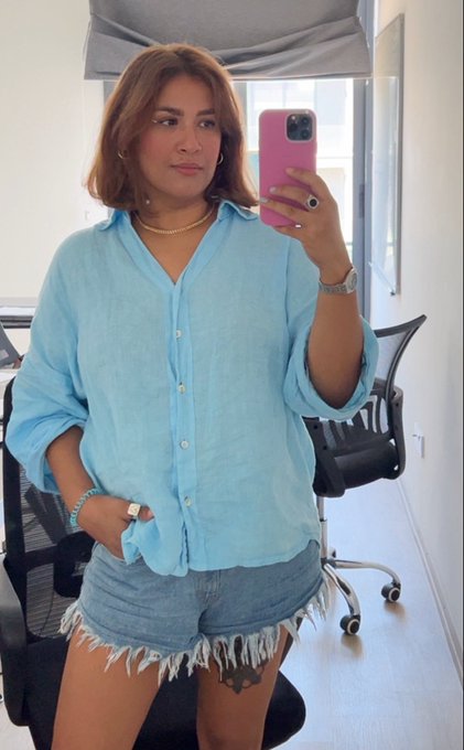A woman with brown hair takes a mirror selfie in a light blue button-down shirt and denim shorts, holding a pink phone.