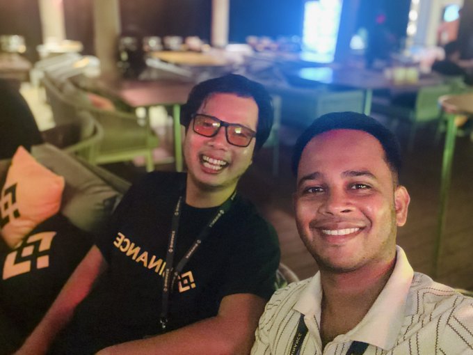 A selfie of two smiling young men at an indoor venue, with one wearing a black Binance t-shirt and glasses, and the other in a white striped polo shirt.
