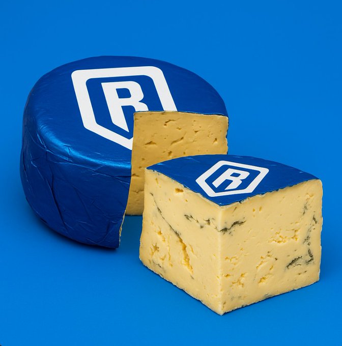 A wheel of blue cheese with an 'R' logo on its rind is displayed next to a cut wedge against a bright blue background.