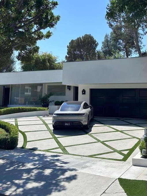 A silver/white Porsche Taycan parked in the driveway of a modern, single-story white home with a black garage door and a geometric concrete driveway featuring grass inlays.