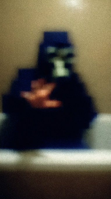A blurry, low-quality photograph showing a figure in dark clothing with what appears to be a helmet or mask with a green glowing element, seated against a beige background.