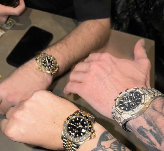 Three friends compare their luxury watches at a table, with a Rolex GMT-Master II, a fully iced-out Audemars Piguet Royal Oak, and a two-tone GMT-Master visible on their wrists. The image of expensive timepieces reinforces the giveaway post's theme of rewarding its crypto community with cash prizes.