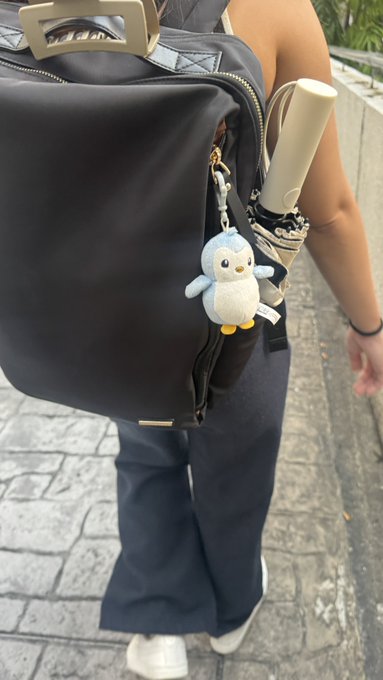 A person walks outdoors carrying a black backpack with a small plush penguin keychain attached, along with what appears to be a folded umbrella.