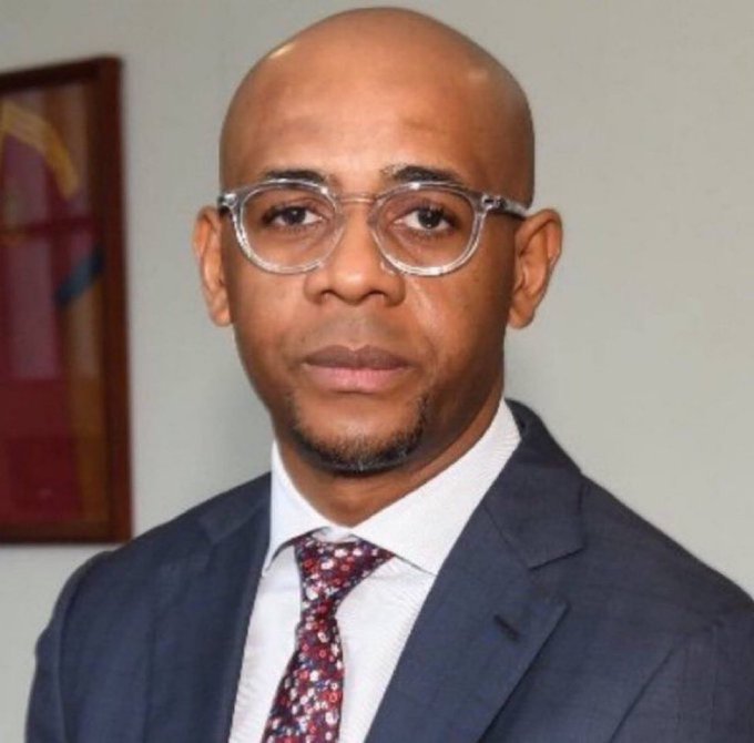 A portrait of a bald man with a goatee wearing clear-framed glasses, a gray suit, white shirt, and a floral patterned tie, posing in front of a wall with a framed picture.