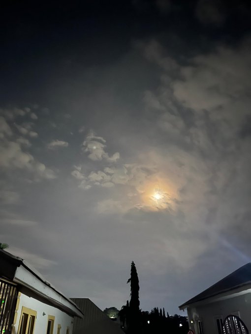 A nighttime photograph of a bright full moon glowing through scattered clouds above silhouetted rooftops and trees.