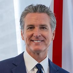 A portrait of Gavin Newsom smiling, wearing a blue suit and tie, with an American flag in the background.