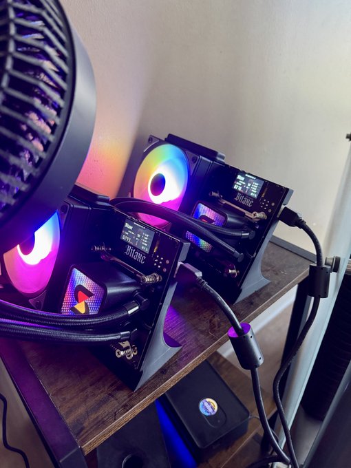 Two Bitaxe Bitcoin miners with vibrant RGB lighting are displayed on a wooden surface, featuring small screens and "Bitaxe" branding. The setup showcases the Bitaxe Gamma Hex, supporting the post's recommendation for at-home Bitcoin mining with these compact, eye-catching devices.