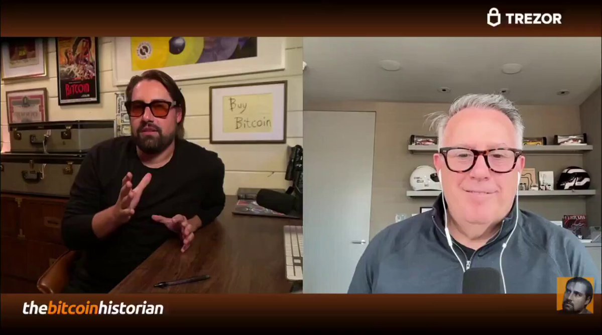 The image shows two men sitting at a wooden desk or table, engaged in an interview-like conversation with each other. They appear to be in professional roles as they hold pens and papers. A man wearing glasses can be seen holding up his cell phone near a picture of himself. The backdrop features various pictures hanging on the wall, which may suggest this setting is a business office or similar environment.