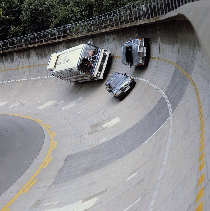 A dramatic scene of three vehicles—a van and two cars—driving on the inside vertical wall of a circular concrete ramp or test track.
