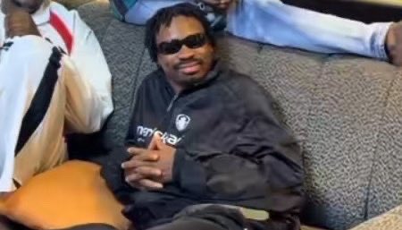 A man wearing sunglasses and a dark tracksuit sits on a couch, with other people visible in the background.