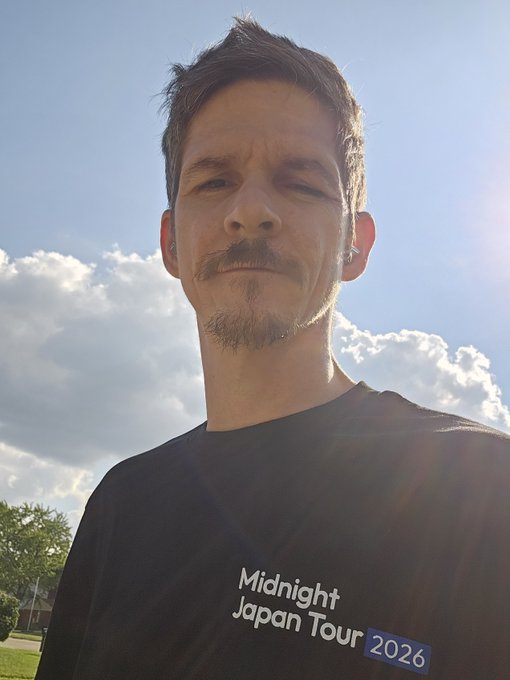 A selfie of a man with a goatee wearing a black t-shirt that reads 'Midnight Japan Tour 2026', taken outdoors against a blue sky with clouds.