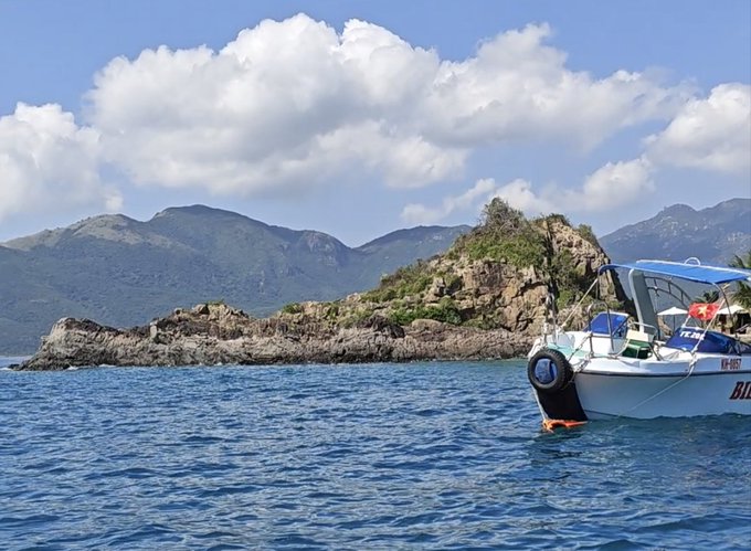 A scenic coastal view showing a white boat anchored in calm blue waters near a rocky island, with mountains and fluffy clouds in the background.
