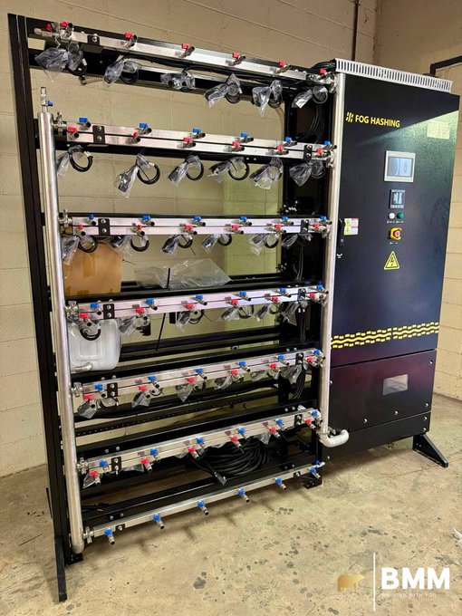 Photograph of a large industrial FOG HASHING liquid cooling system with multiple rows of black tubing and red/blue valves mounted on a metal frame, branded with the BMM logo in the corner.