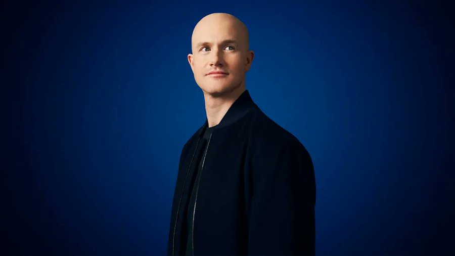 A bald man in a dark jacket poses against a solid blue background.
