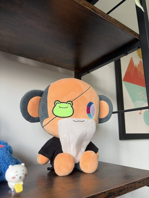 A Chimpi plush toy sits on a dark wooden shelf, featuring an orange monkey-like character with a green frog face patch and an "x" over one eye, matching the post's teaser of a "New Chimpi plush loading" for Wise Beard Wednesday in the Chimpverse.