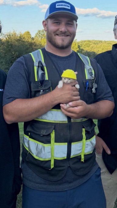 A smiling man in a Komatsu cap and reflective safety vest holding a small kitten wearing a yellow construction hard hat.
