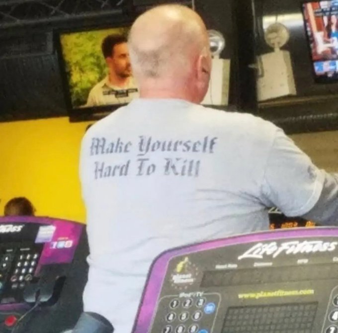 A bald man wearing a gray t-shirt that reads "Make Yourself Hard To Kill" in Old English font sits at a Life Fitness cardio machine, with multiple TV screens visible in the background, one showing what appears to be a sports broadcast.