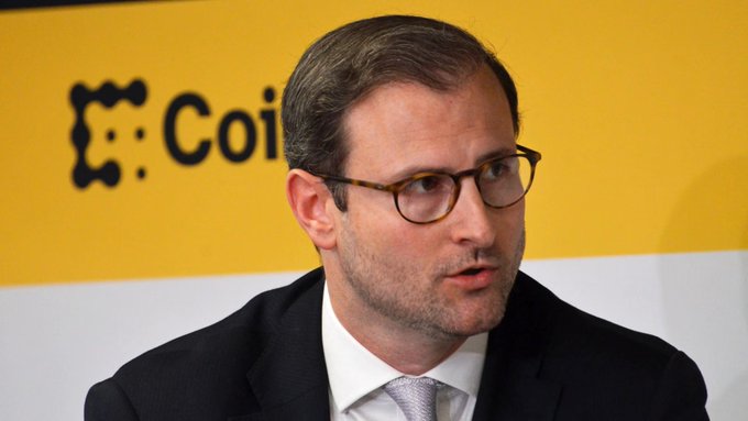 A man in glasses, dark suit, and tie speaks at an event with a yellow and white background displaying partial "Coi" text. The image relates to the post as it depicts White House crypto adviser Patrick Witt discussing his optimistic outlook on the CLARITY Act ahead of its Senate cloture vote.