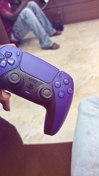 A hand holds a purple-and-black PlayStation DualSense controller, with seated people visible in the blurred background.