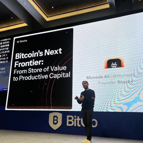 A man giving a presentation on stage in front of a large screen displaying 'Bitcoin's Next Frontier: From Store of Value to Productive Capital' by Muneeb Ali, Founder Stacks.