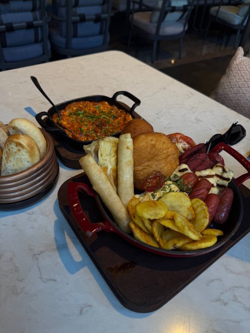 A photo of a restaurant meal featuring a cast-iron skillet with red handles filled with sausages, fried items, and chips, alongside a smaller pan of stew and a basket of bread.