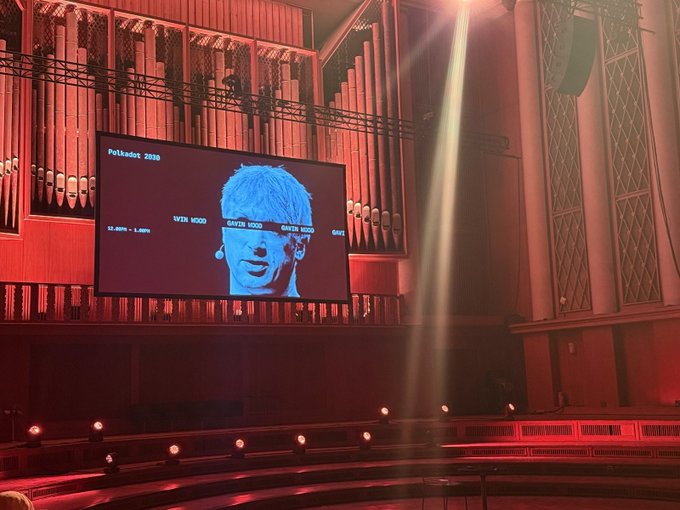 The image shows a large screen displaying a man's face with headphones around his neck. The screen also has text that reads "Web3 isn't just Web2 plus crypto—it's a fundamental shift in how we build At its heart is the @Polkadot Trinity: the convergence of scalability, interoperability, and user abstraction to make Web3 invisible." This image seems to be related to a talk given at the Web3 Summit.