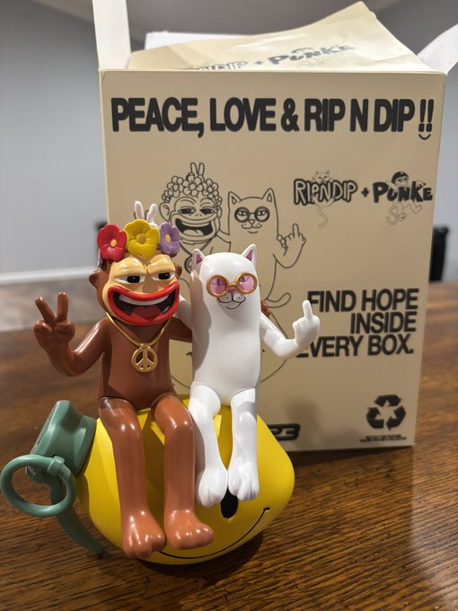 A RIPNDIP 'Peace, Love & Rip N Dip' blind box containing a figurine of the Nermal cat and Lord Nermal monkey character sitting on a smiley bomb.