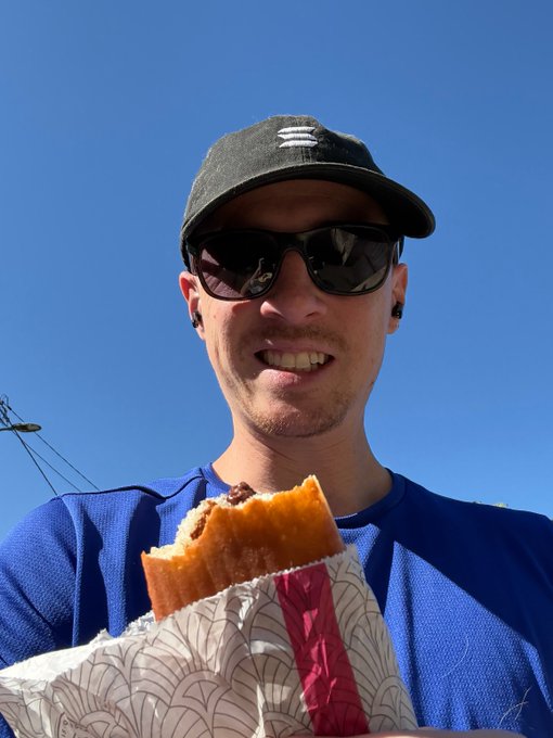 A selfie of a smiling man in a black cap, sunglasses, and blue athletic shirt, holding a partially eaten pastry against a clear blue sky.