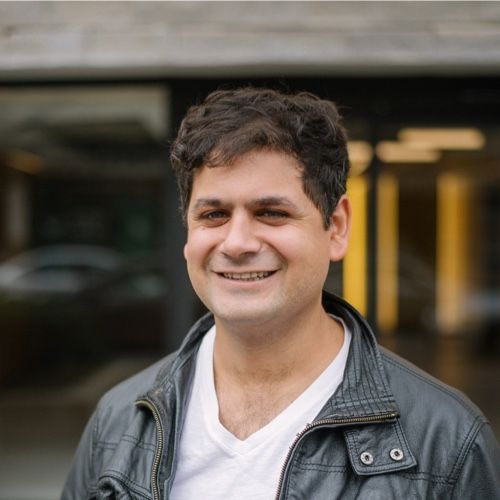 A portrait photograph of a smiling middle-aged man with curly dark hair, wearing a white V-neck shirt and a gray leather jacket, standing outdoors.