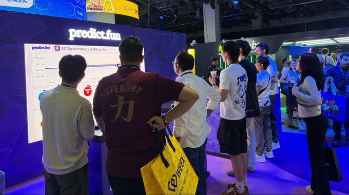A group of attendees gathers around a brightly lit "predict.fun" booth at what appears to be a conference, with a leaderboard displayed on the screen and someone wearing a "Superdry" shirt holding a yellow "PYWER" bag in the foreground. The scene captures user engagement with the predict.fun platform, likely showcasing its prediction market activity during the event.