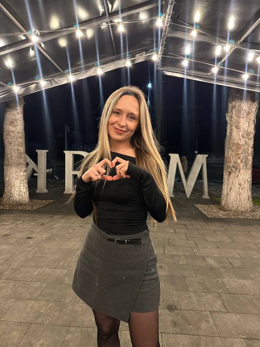 A blonde woman making a heart shape with her hands while posing in front of large decorative letters under string lights at night.