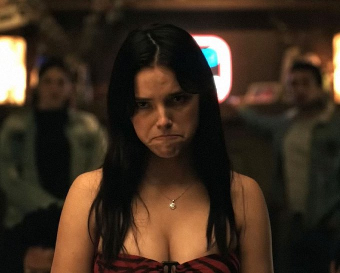 Cinematic still of a young woman with dark hair wearing a red top, frowning in a dimly lit indoor setting with blurred figures in the background.