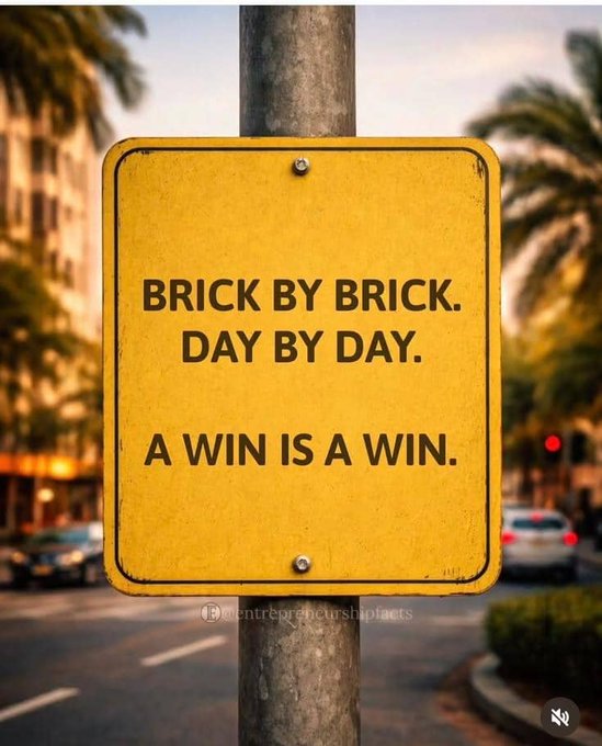 A yellow street sign on a pole reading 'BRICK BY BRICK. DAY BY DAY. A WIN IS A WIN.' with palm trees and a street scene in the blurred background.