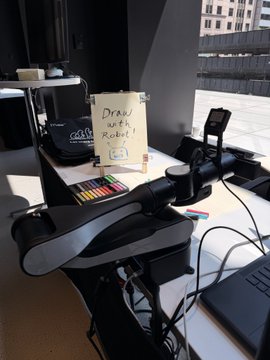 A robot arm operates at a demonstration table displaying a handwritten sign reading "Draw with Robot" in multiple colors, alongside a box of colorful markers. The setup is positioned near a window at Willis Tower, where attendees of the Web3 Investor Day event can control the robot to create their own drawings.