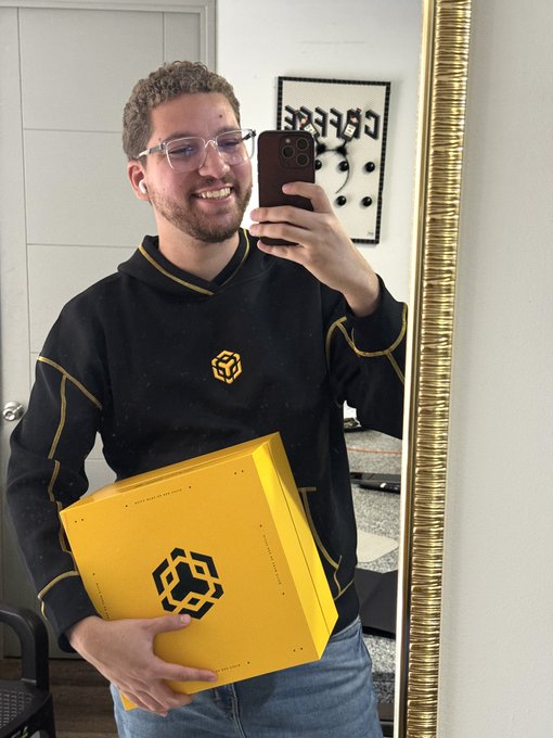 The image features a man holding a yellow box with a black logo, possibly containing a new item or gift for someone else. The man appears to be taking a selfie using his cell phone while holding the box. The background of the photo includes a mirror that reflects the room and another person sitting in the background.