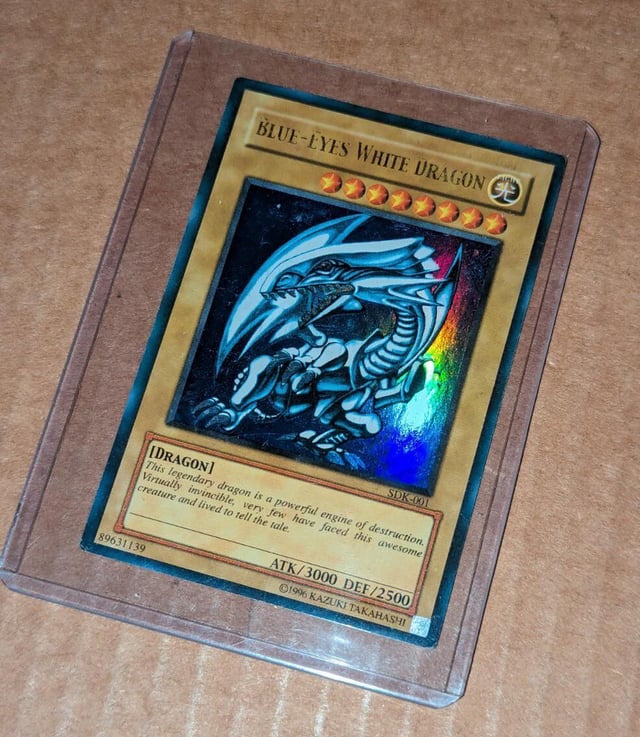 The image features a Pokemon trading card, specifically a Blue-Eyed White Dragon card, placed inside a clear plastic sleeve. The card has a gold background with a dragon illustration and text that reads "Blue Eyes White Dragon". The card is encased in a clear plastic sleeve, which is resting on a brown surface.

The post also mentions the collector's name as "Algorand Collector" and their favorite Pokemon being Blue-Eyed White Dragons.