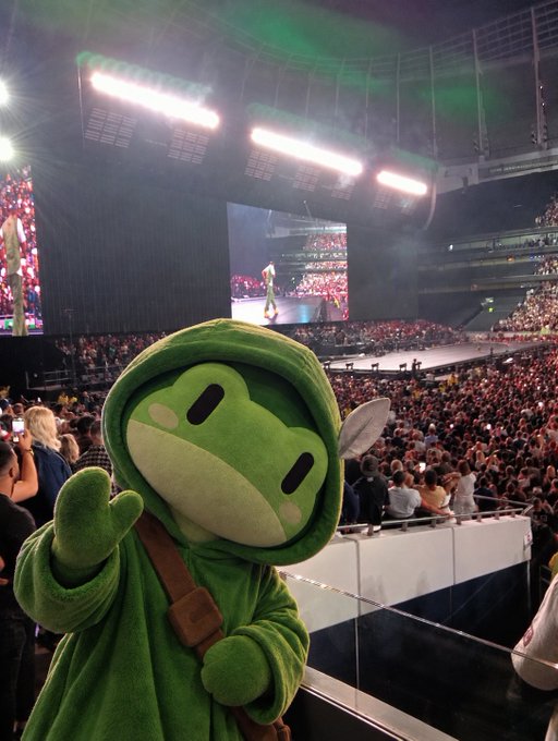 A green frog mascot character in a hoodie waving at the camera in front of a large arena concert crowd watching a performer on screen.