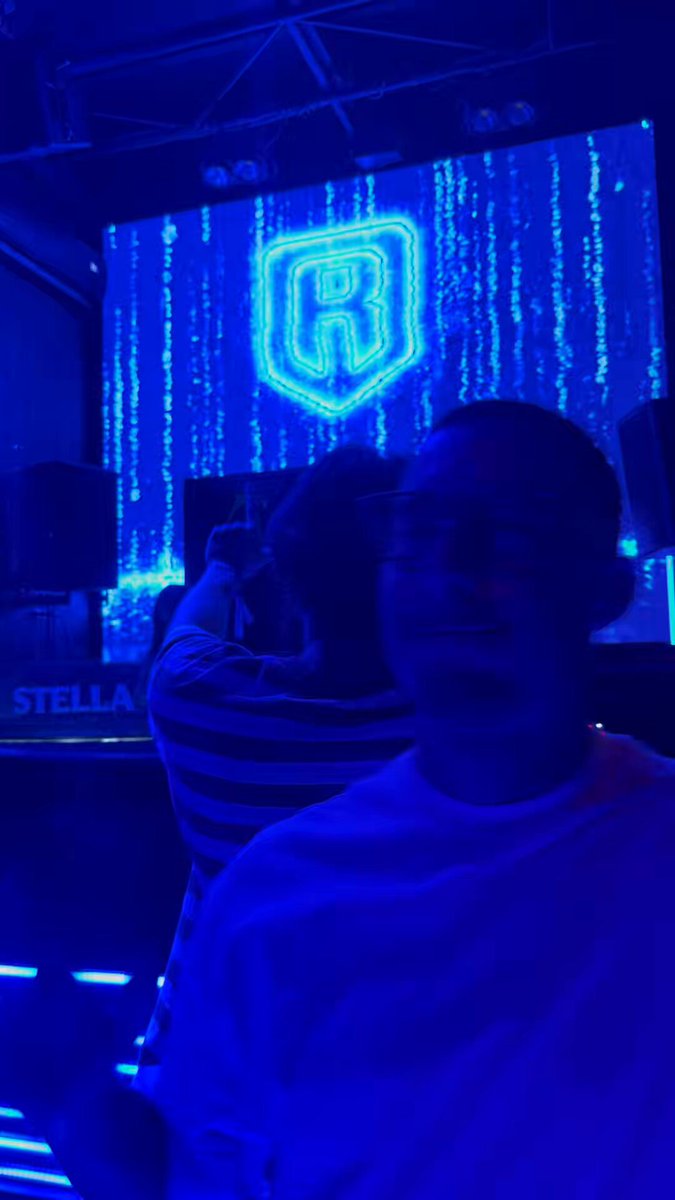 The image features a man wearing glasses, standing next to a neon blue sign that says "R". He appears to be taking a picture of himself with his phone. The background includes speakers and lights, suggesting an indoor setting like a club or concert venue. The caption for the image reads "Que viva Ronin y Kanstar! The Matrix is here @Ronin_Network @KanstarWorld".