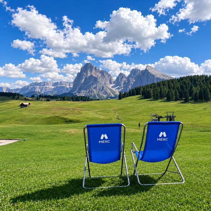 Two blue folding chairs branded with the "MEXC" logo face a stunning alpine landscape of jagged mountain peaks under a partly cloudy sky, embodying the post's "trading setup with a view" by trading the desk for a scenic meadow retreat—rated highly on the view, if not on the Wi-Fi.