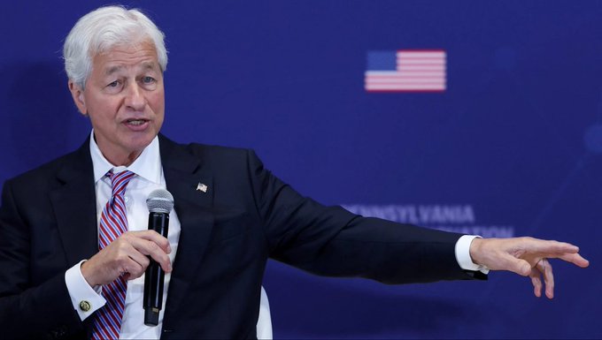 A man with white hair in a dark suit speaks into a microphone while gesturing, standing in front of a blue backdrop with an American flag and the word "PENNSYLVANIA" partially visible.