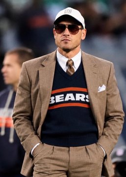 A man stands on a sports field wearing a vintage Chicago Bears sweater, tan suit, tie, sunglasses, and a white cap.