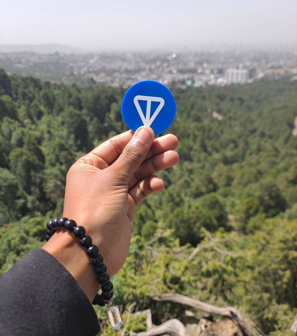 The image features a person holding up a blue sticker with a white triangle symbol on it. This sticker has the word "Telegram" written on it. The person's hand is visible above their wrist, while the sticker is held at an angle in front of them. The background shows a cityscape or hillside view, suggesting that this scene takes place outdoors.